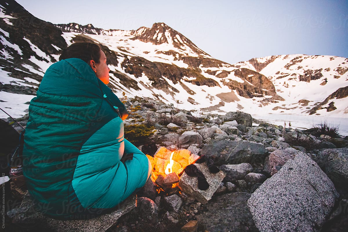 A young woman snuggled in a sleeping bag sitting beside a small fire in the mountains