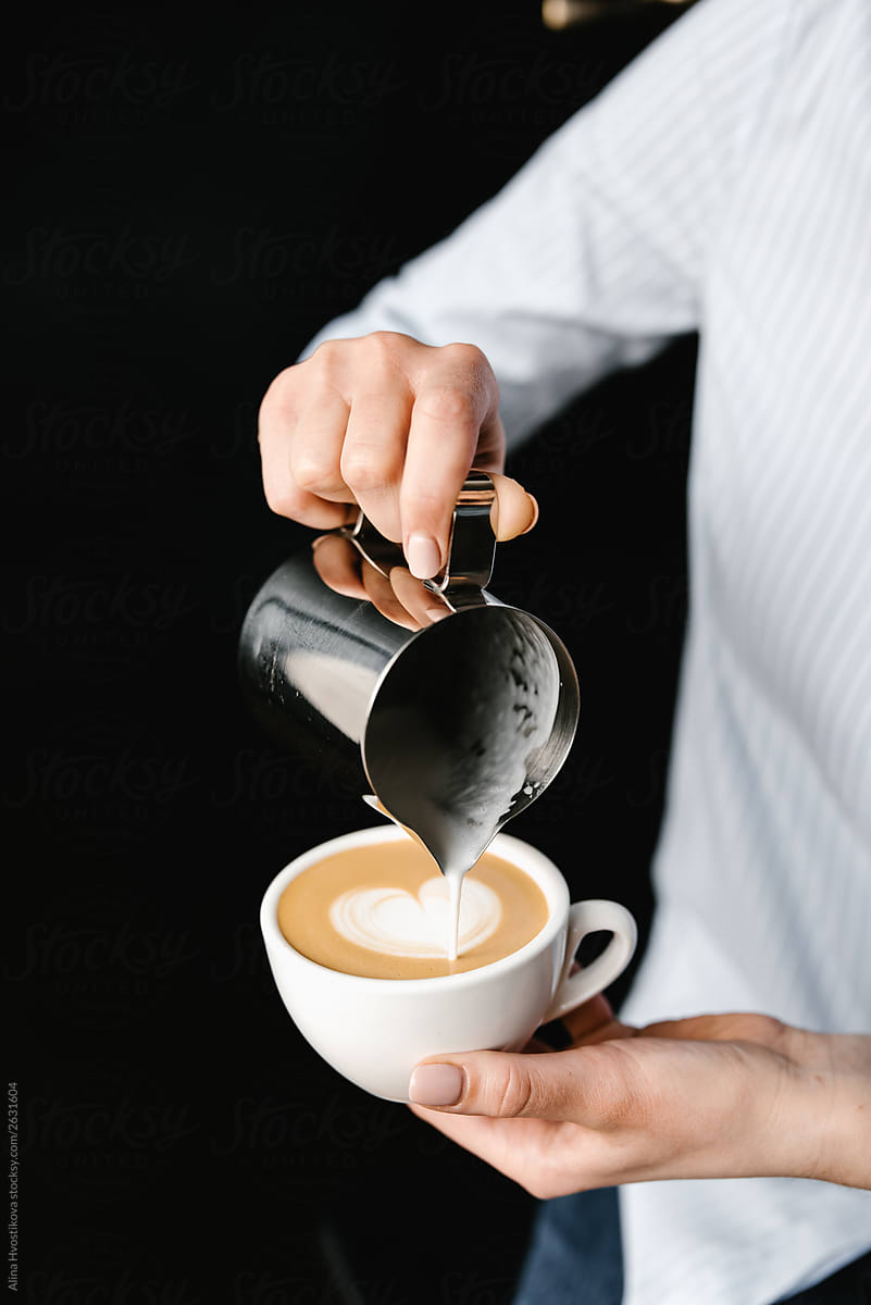 Female pouring creamy milk in coffee cup from iron pitcher.