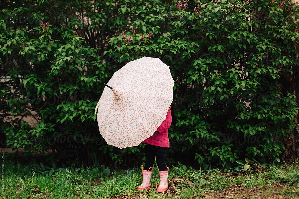 Toddler girl in a pink jacket and rain boots outside with an umbrella.