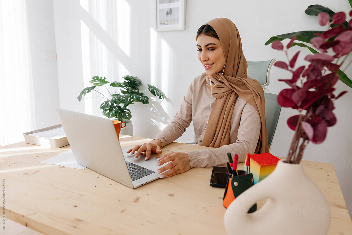 Positive woman in hijab working on laptop in workplace