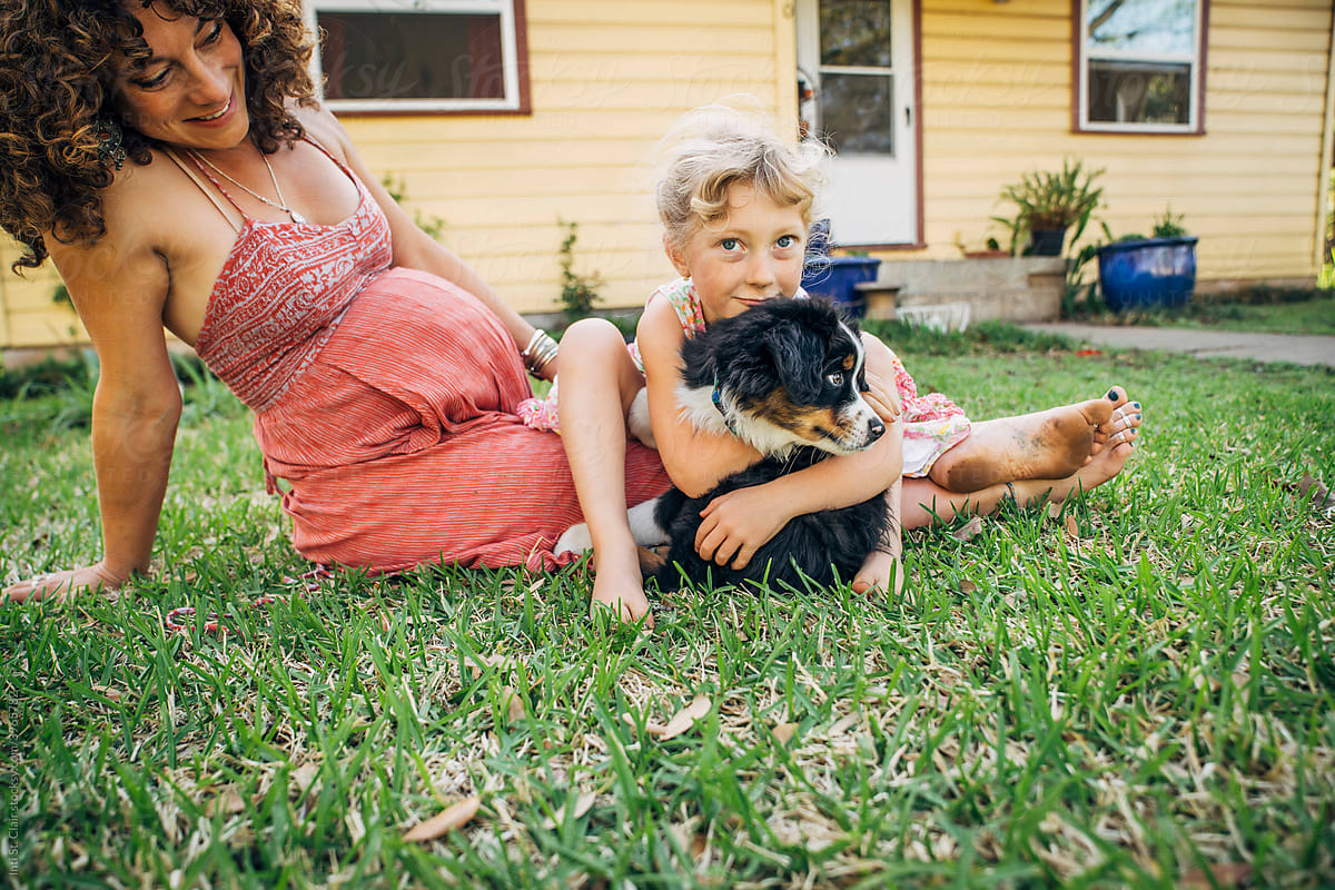 Girl sitting on pregnant mom\'s lap hugging puppy in yard