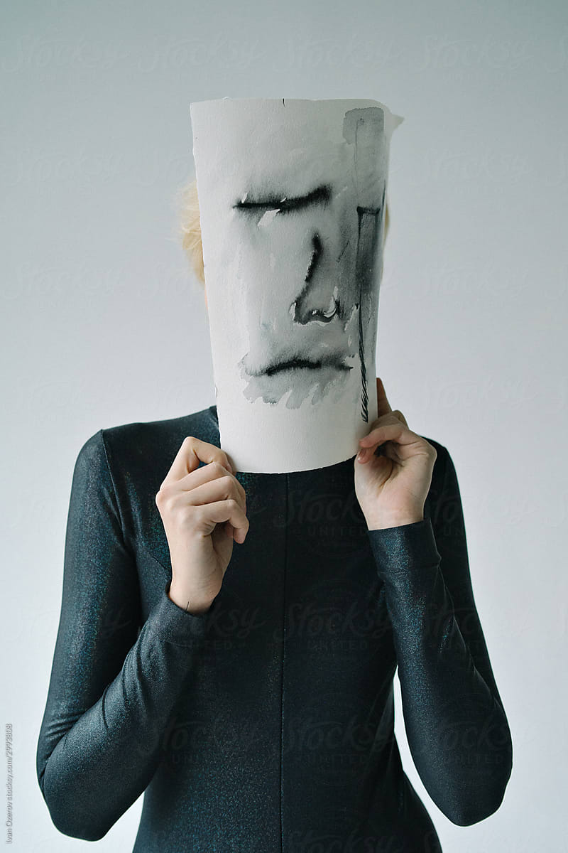 a slender girl stands covering her face with a sheet of paper with a sad face depicted on it