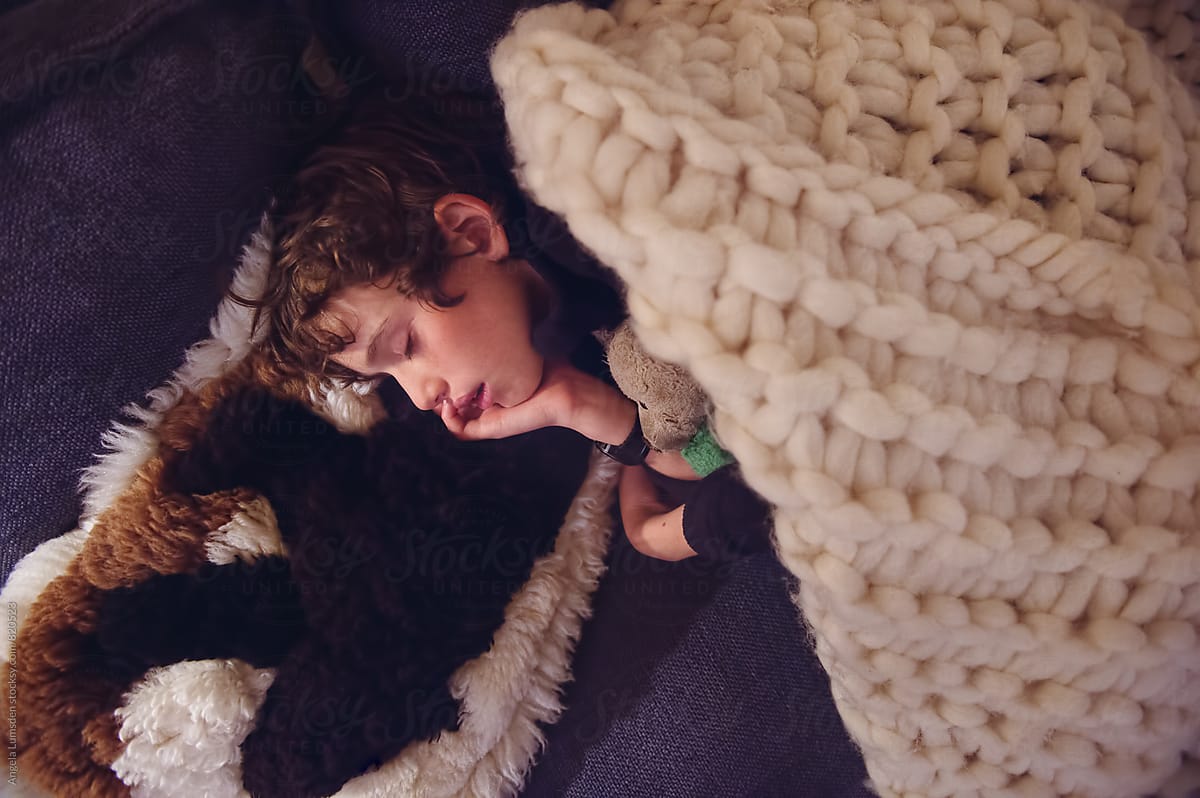 Boy asleep on a couch with a teddy bear under a large knit blanket