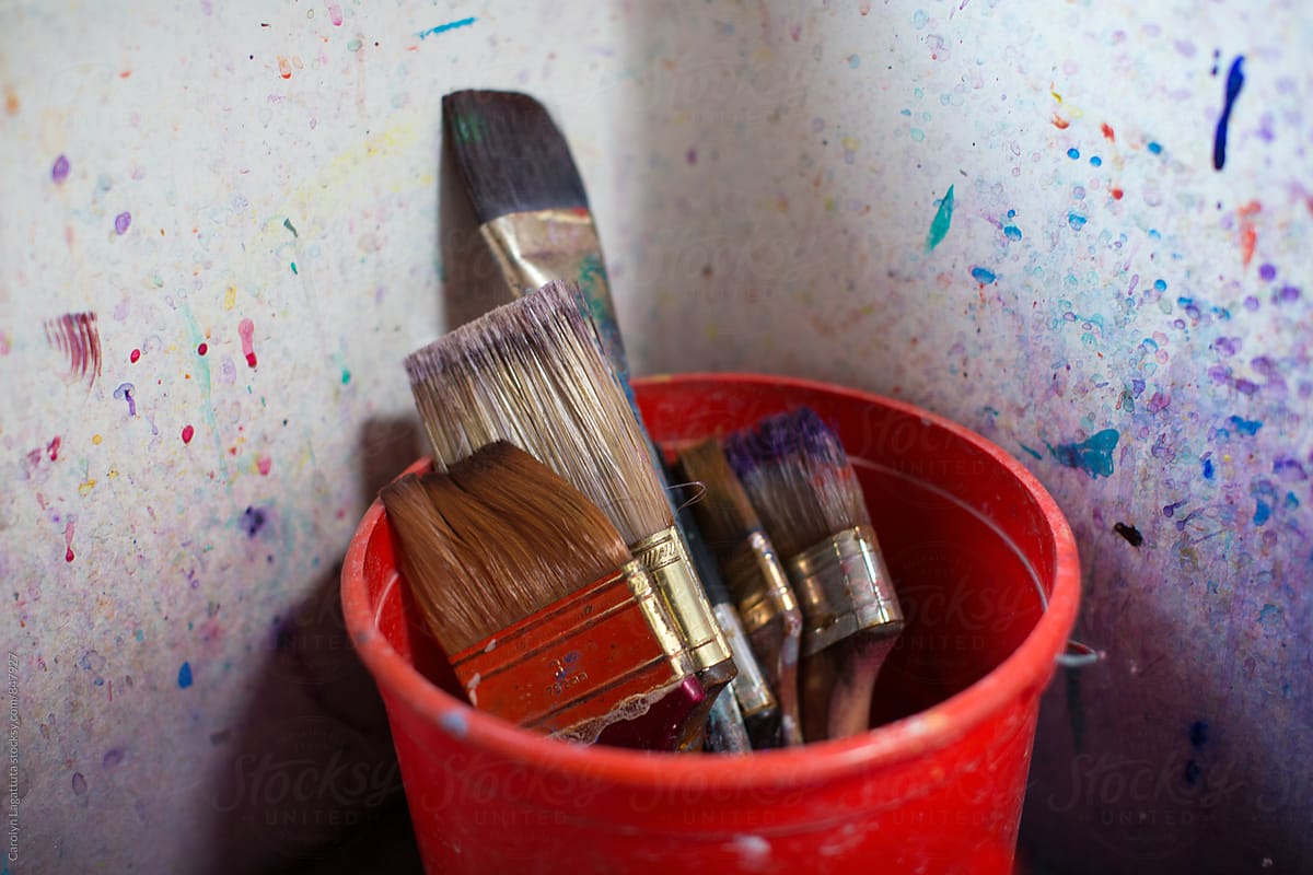 Many paint brushed in a red bucket inside a paint splattered sink