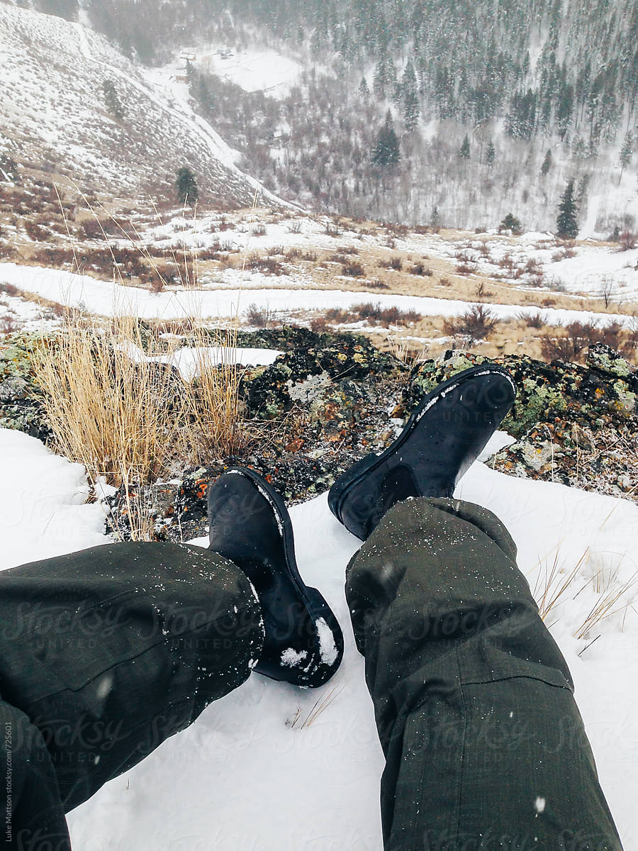 Man In Green Snow Pants And Black Boots Sits On Top Of Hill Covered In Snow