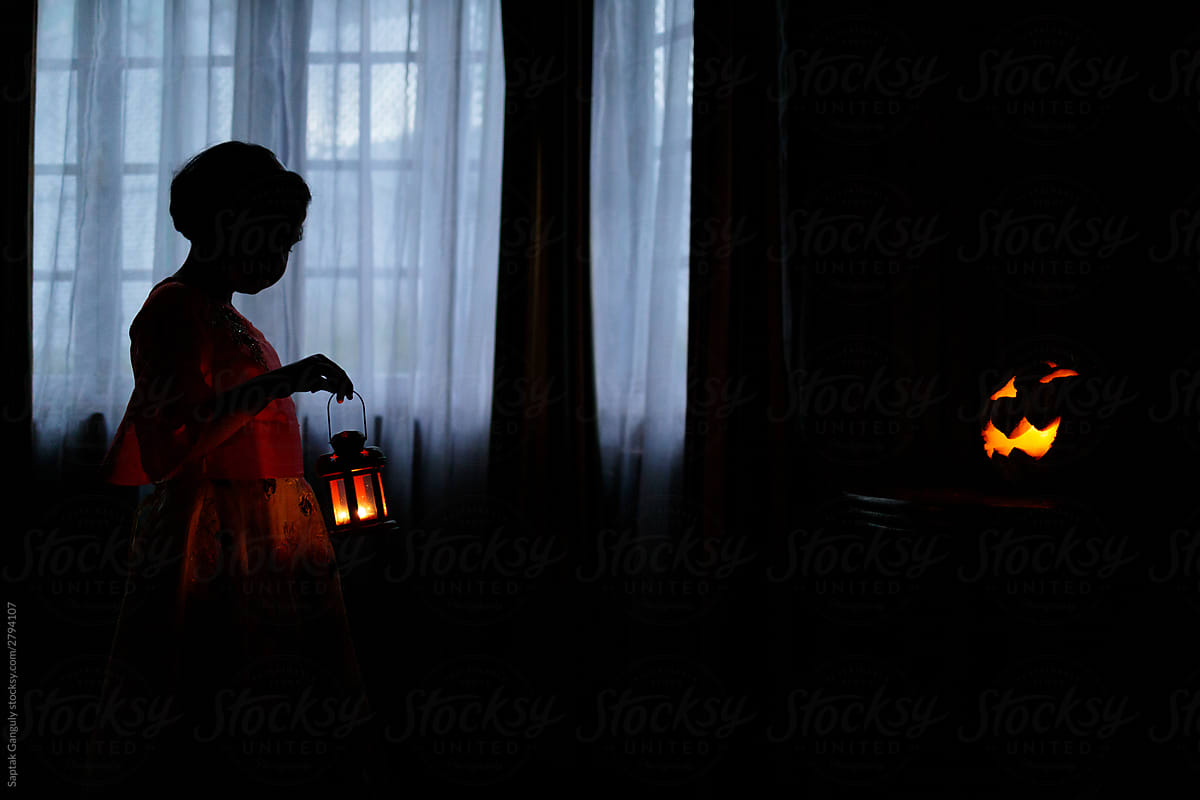 Silhouette of a little girl with lantern walking towards a jack-o'-lantern at dusk