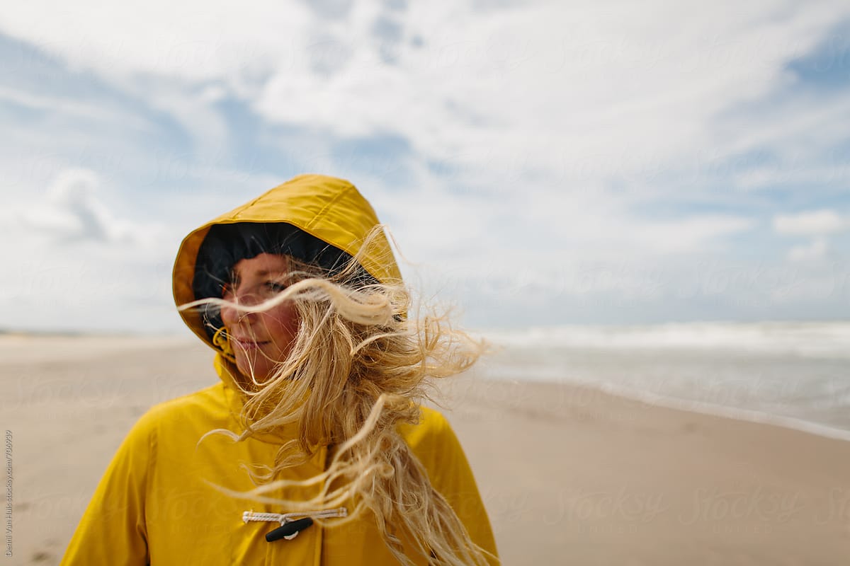 Woman On A Windy Beach Wearing A Yellow Raincoat With Hair Blowing Everywhere By Stocksy 6759
