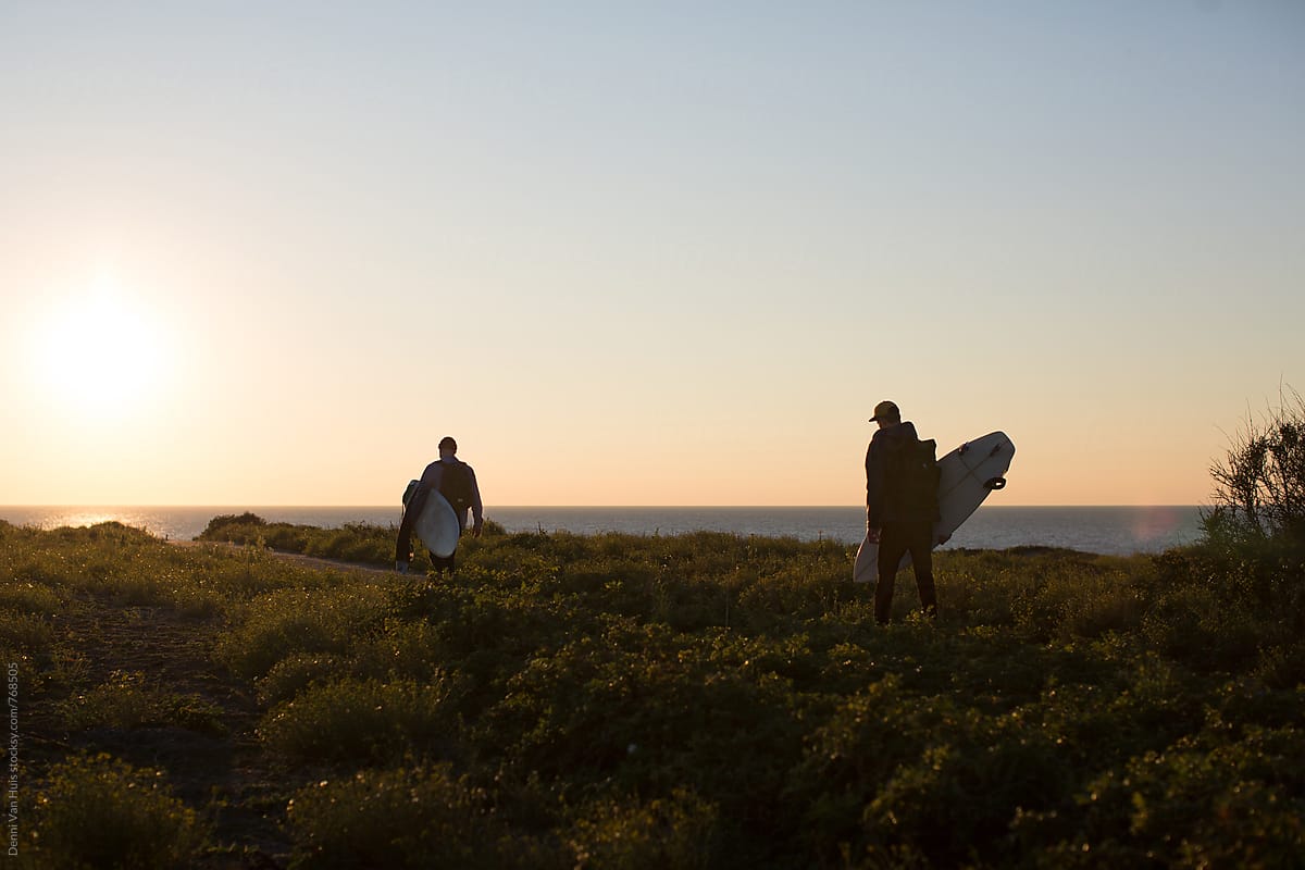 Silhouette of two friends hike through the dunes with surfboards in search of waves.