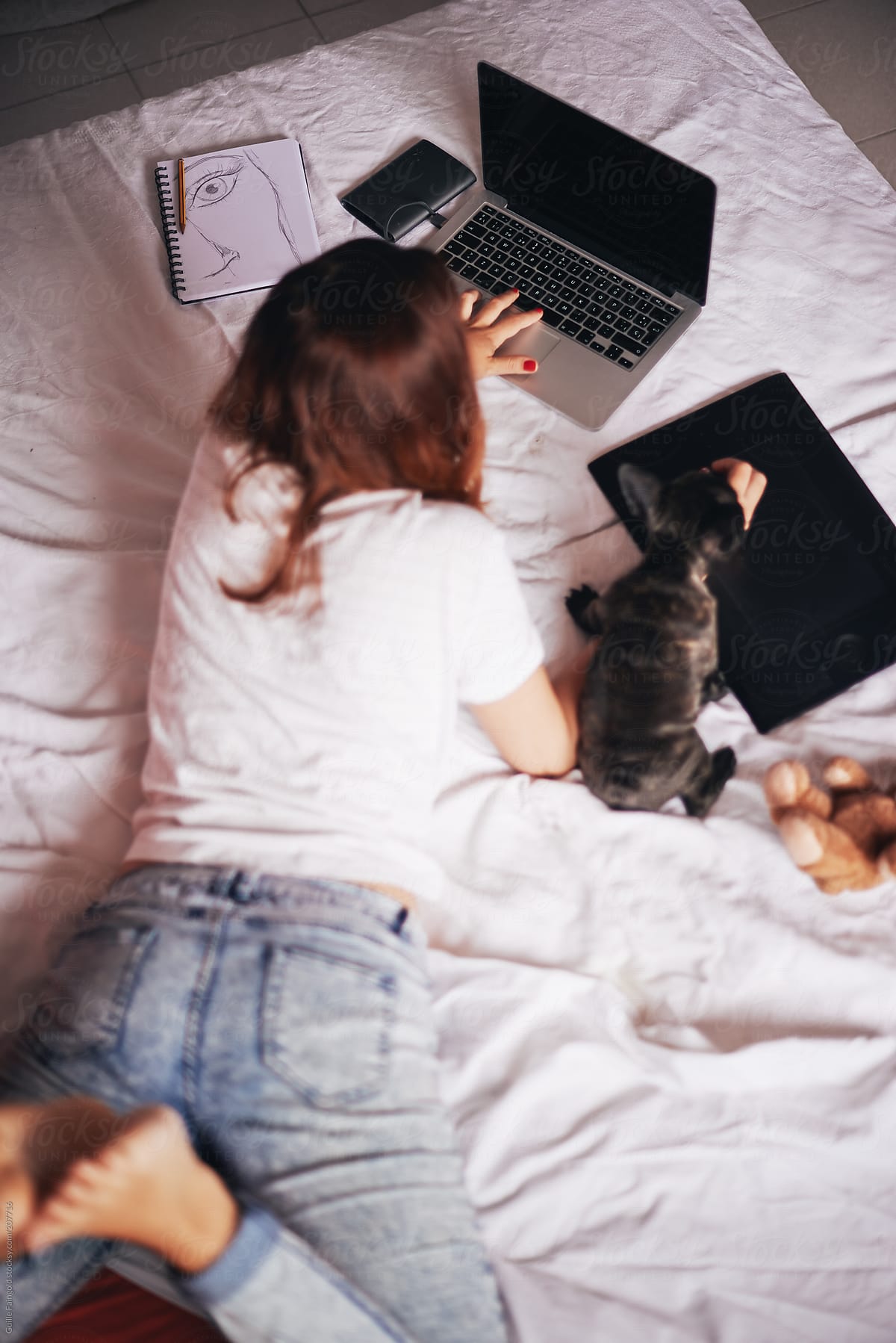 Girl typing on laptop and playing with puppy on bed