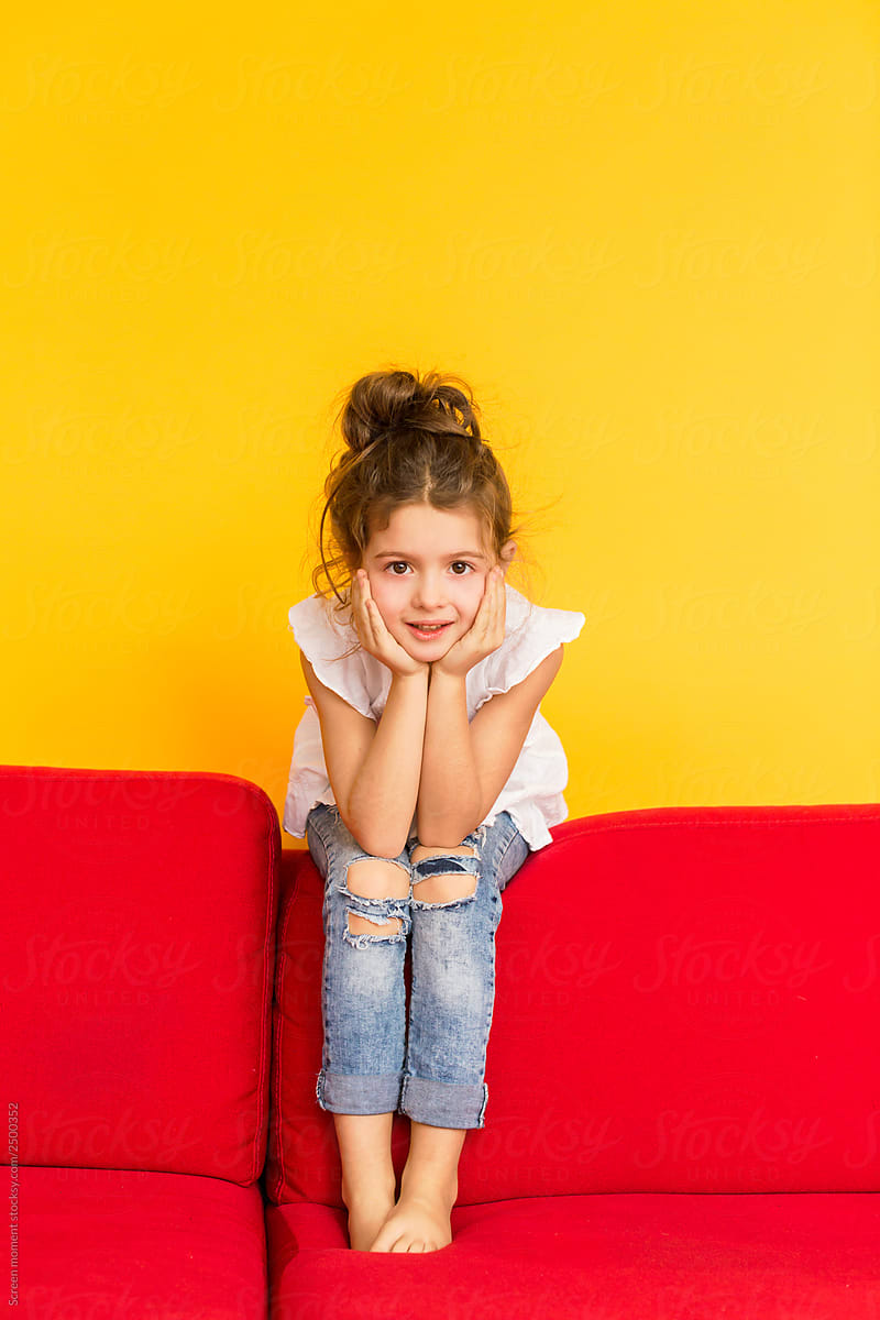 Little cute girl with dark hair in a white blouse and jeans sits on a red sofa on a yellow background. Little girl grimaces