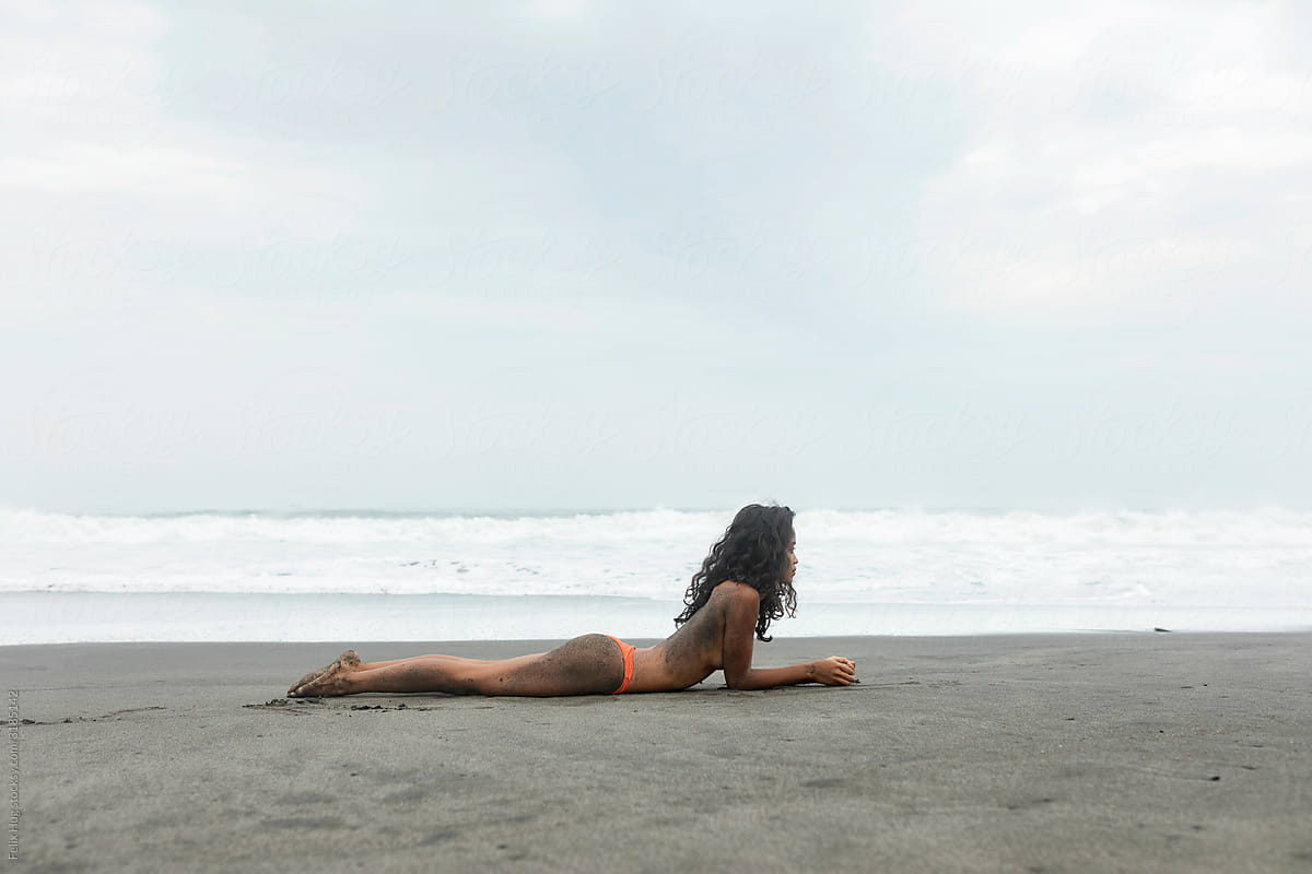 A topless dark girl with red bikini bottoms is lying belly down o a volcanic dark beach in Bali