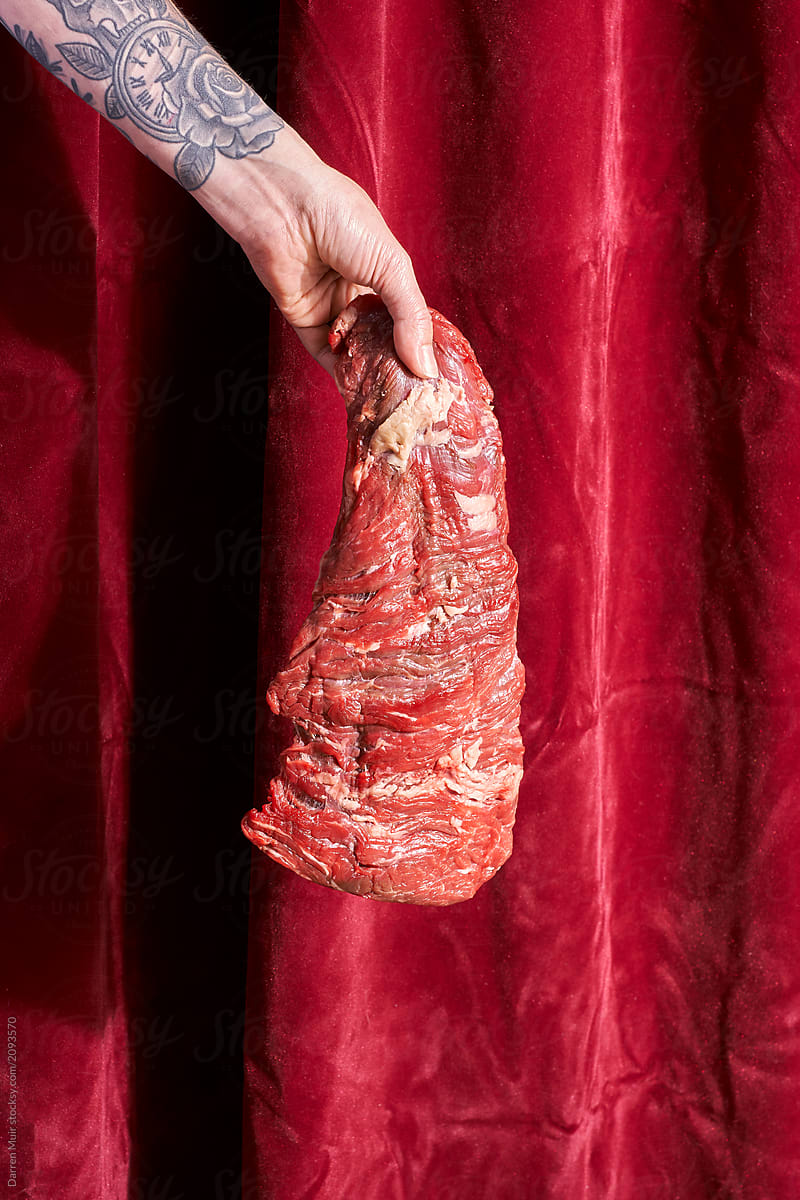 Slab of red meat being held by a hand against a crushed red velvet curtain....