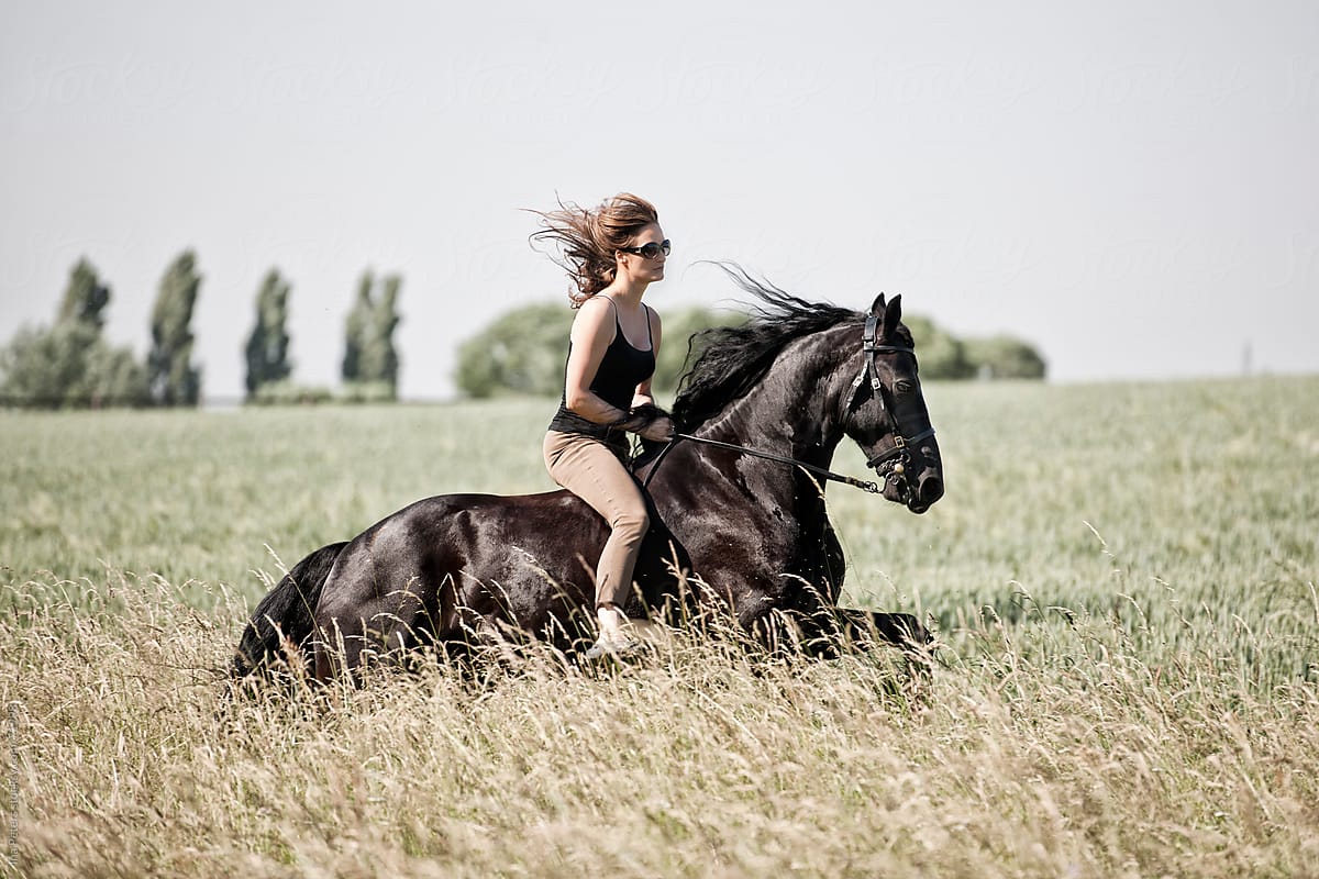 People: Woman Riding A Black Horse In A Agrass Meadow by Ina