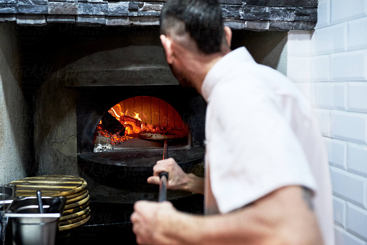 chef in uniform putting pizza into wood fired oven.