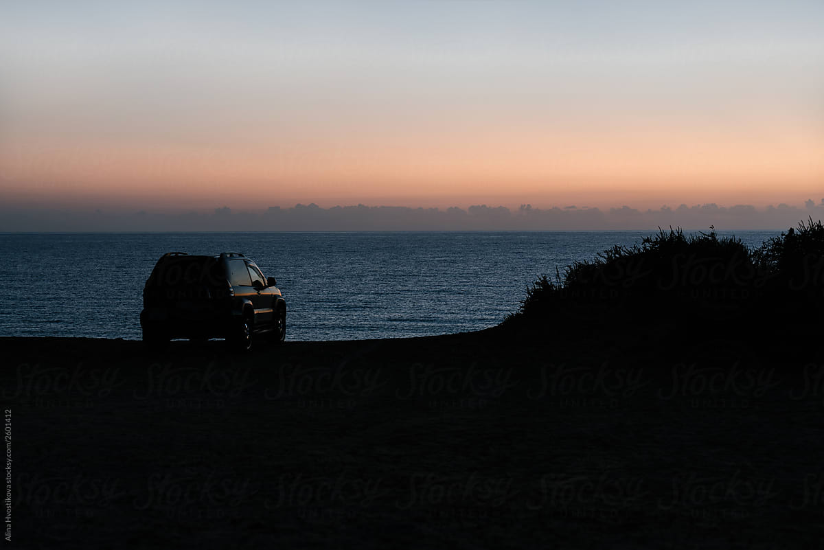 Car standing over cliff of shore near ocean after sunset.