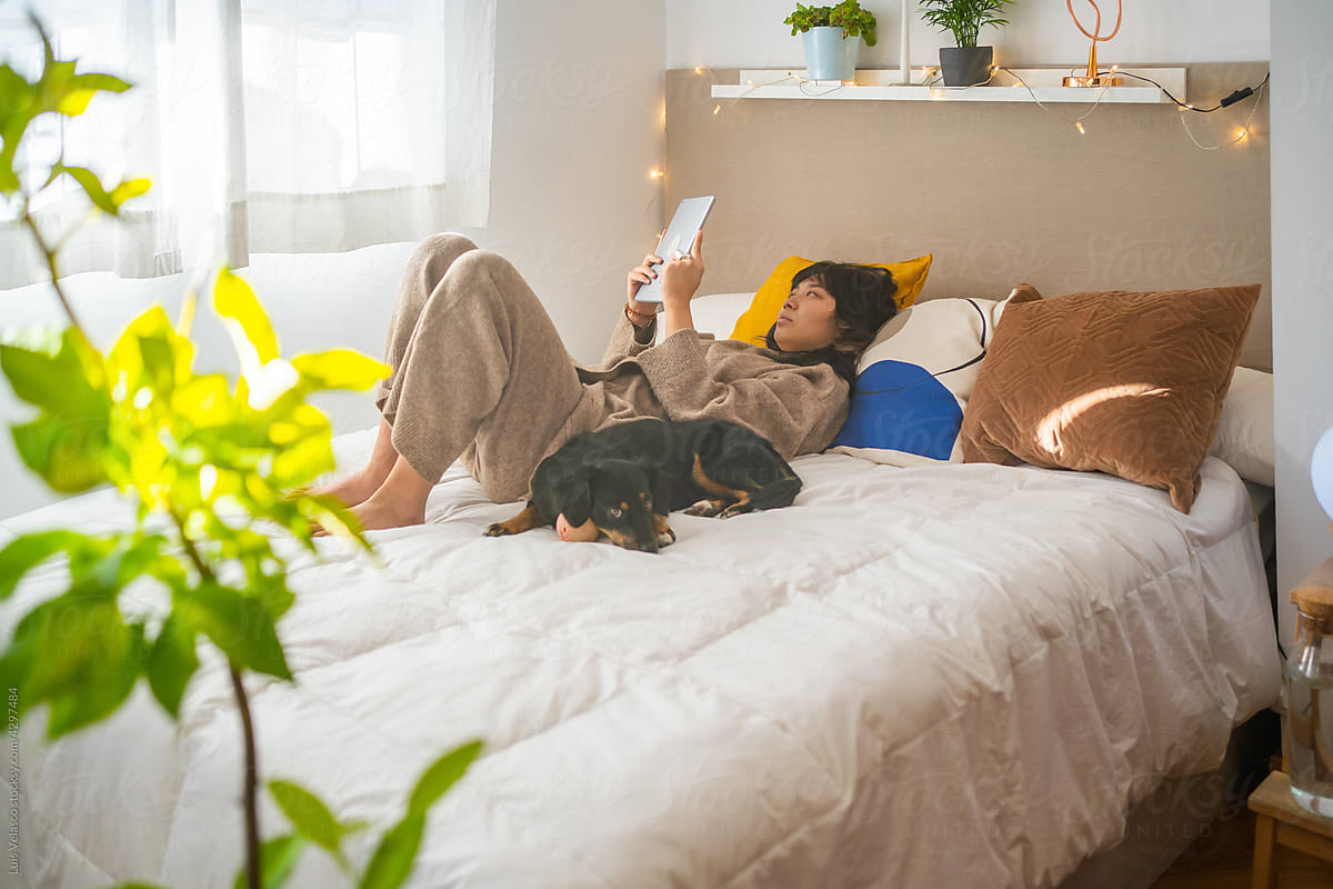 Asian Woman Laying On The Bed And Reading With Her Dog.