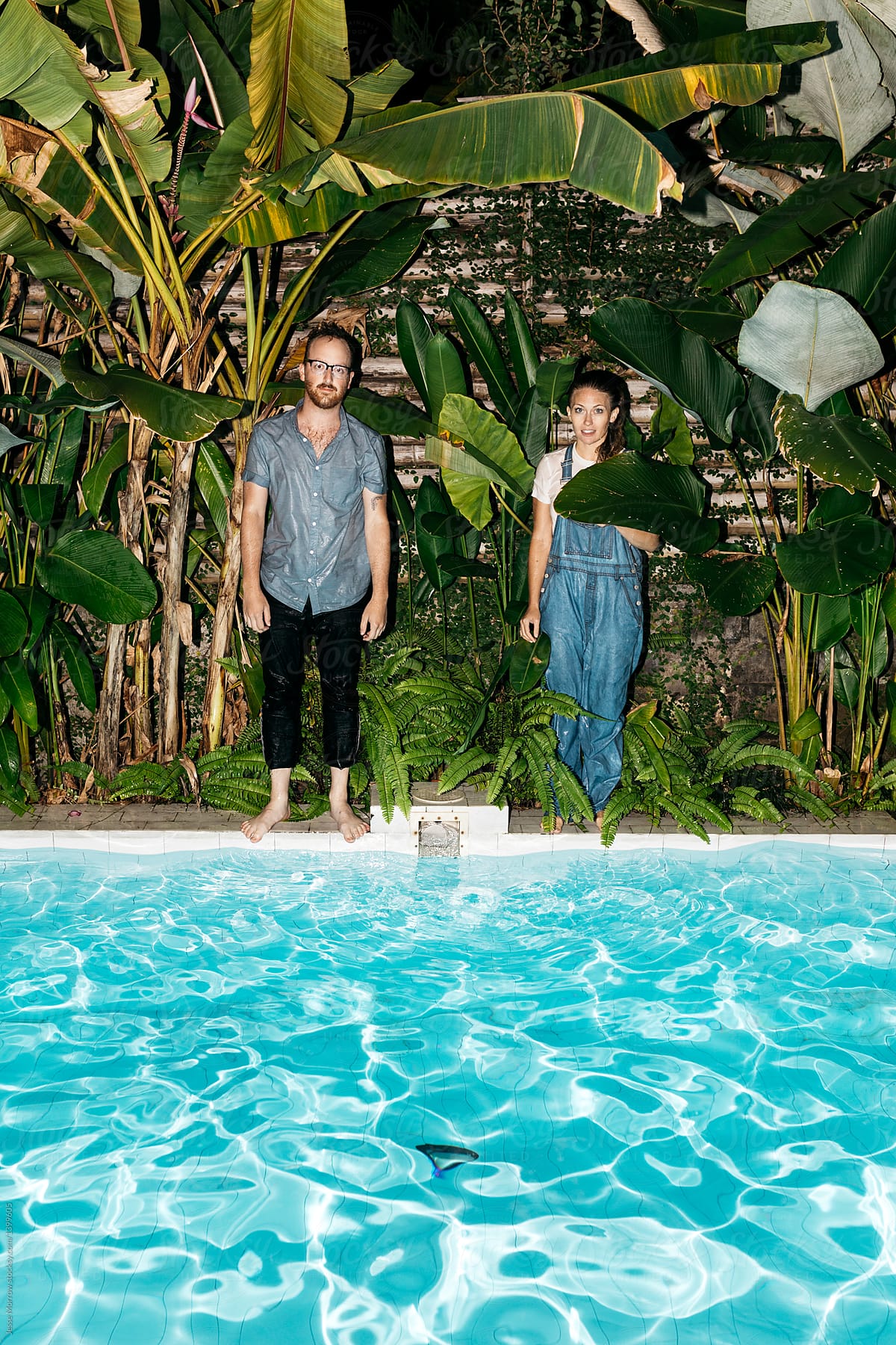 Young couple standing in tall green foliage jungle next to pool in tropical night time swim