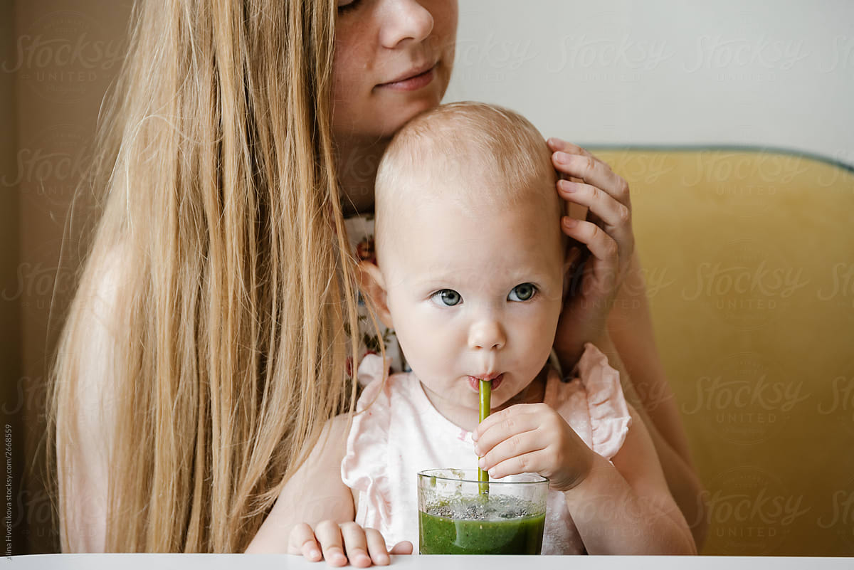 Baby girl with blue eyes drinking green smoothie with mom.