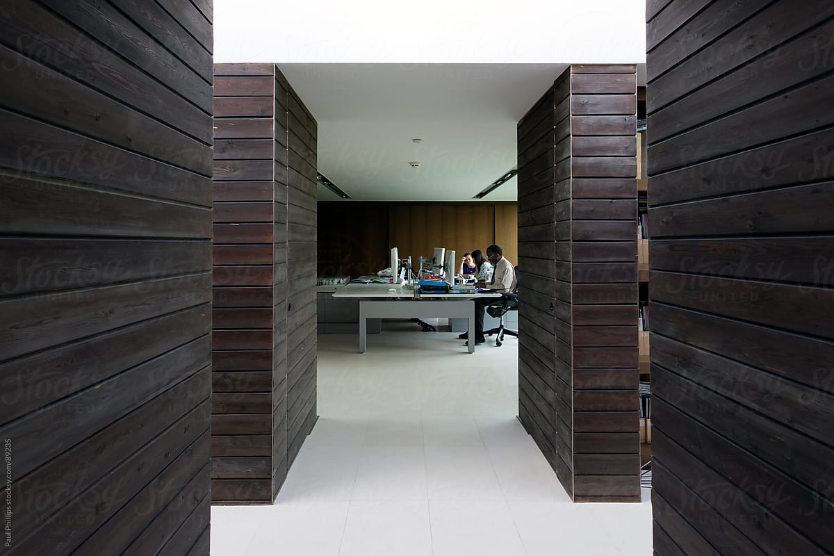 Interior of modern office with wooden corridors leading to working area. Three people working