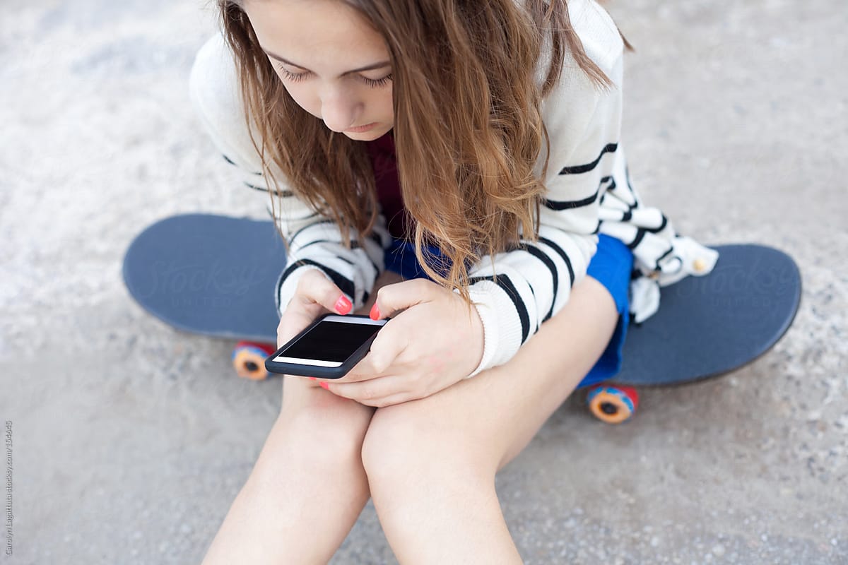 Teen girl sitting on her skateboard and playing with her cell phone