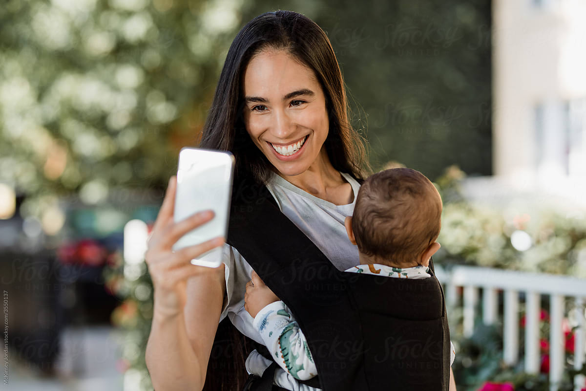 Happy Woman Uses Smartphone While Carrying Baby