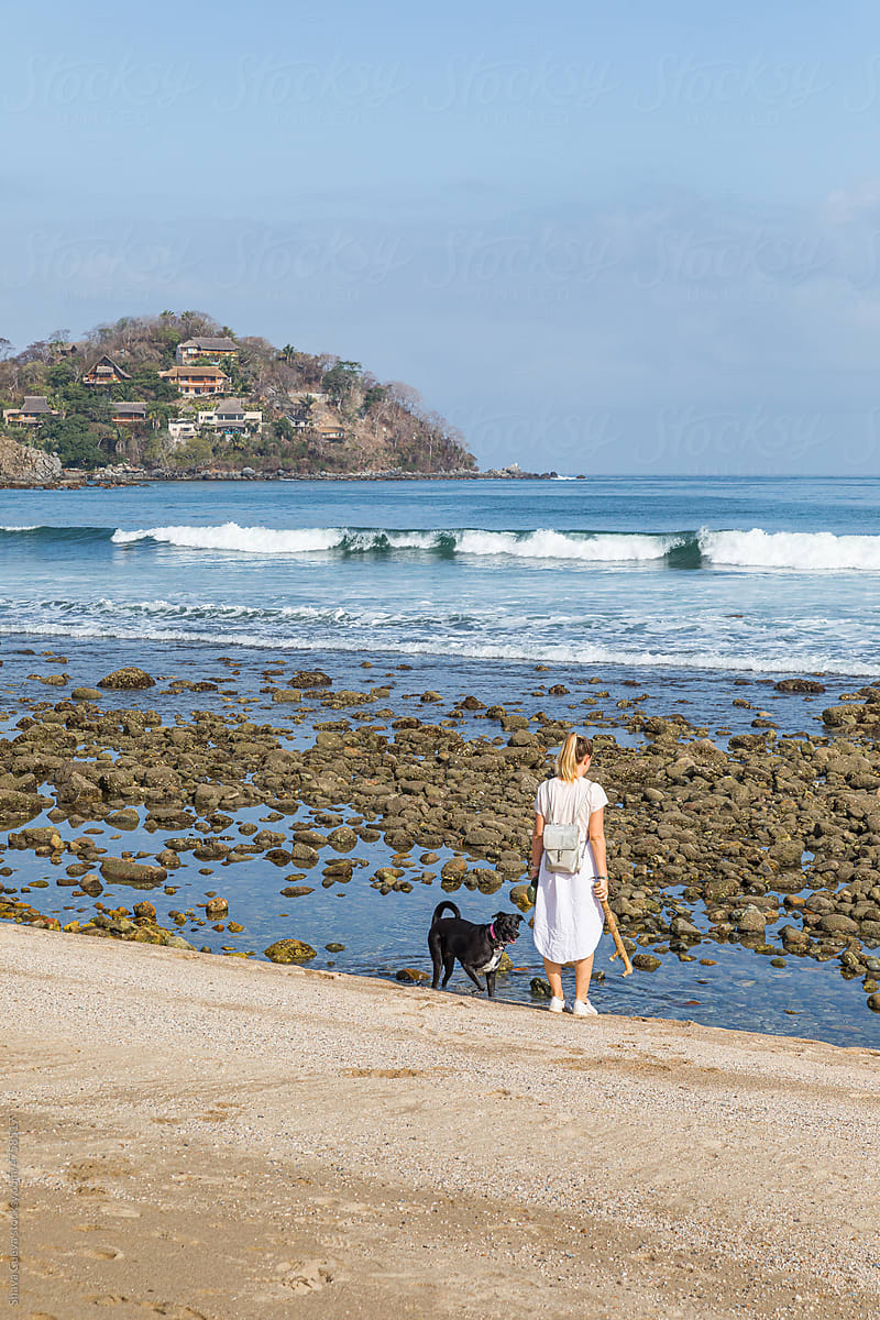 Woman in white clothes with her dog in front of a beach with rocks