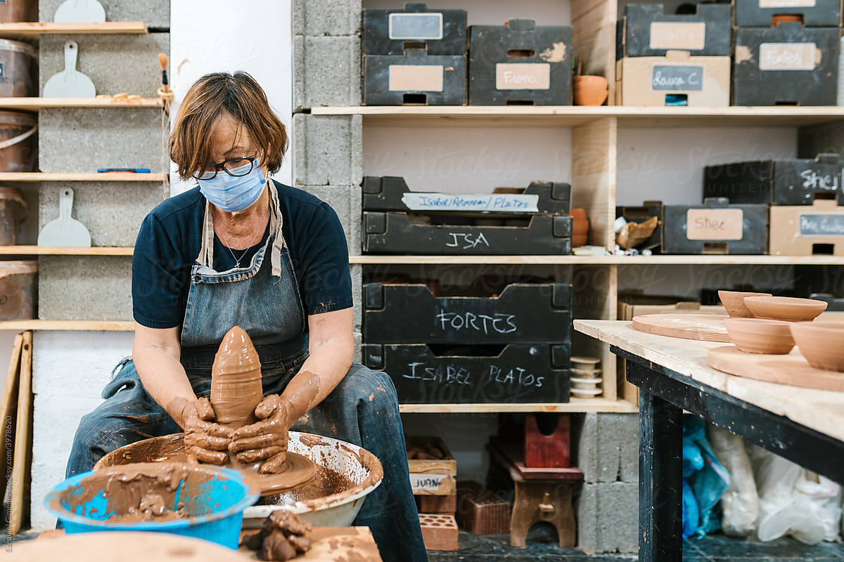 Mature craftswoman making clay vase near shelves
