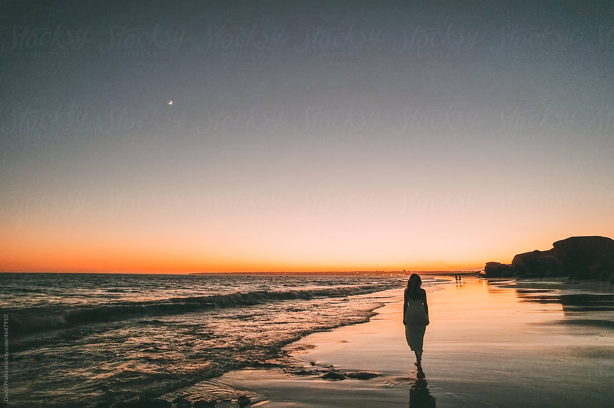 Barefooted lady standing on sandy seashore at sunset