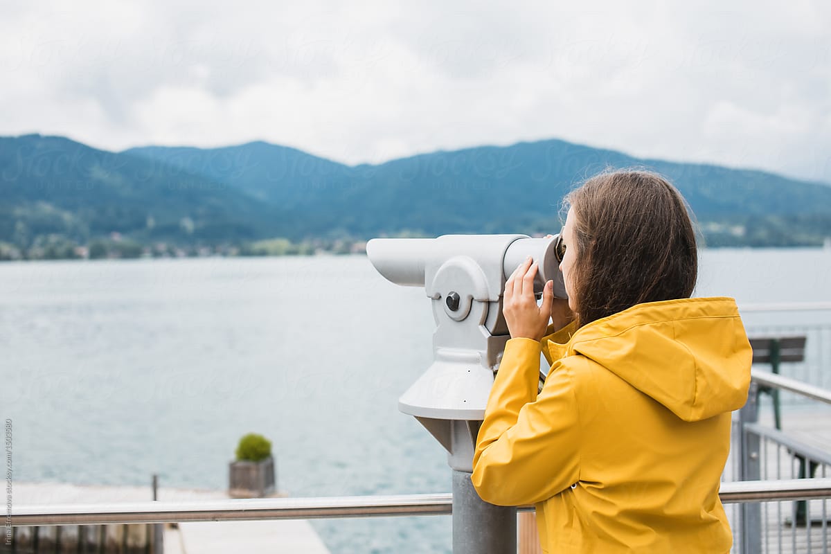 Girl in a yellow raincoat looking through coin-operated telescope at the opposite bank of the lake in Alps
