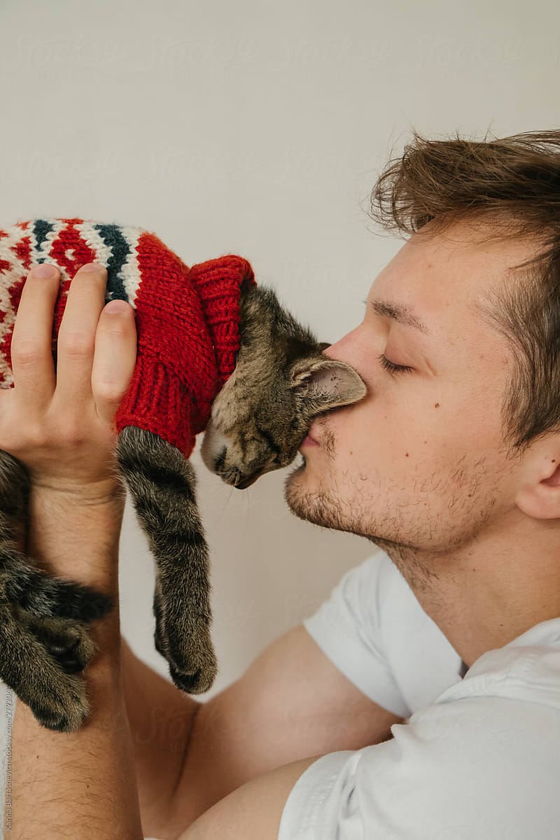 guy in a white T-shirt kisses a kitten in a red sweater on his forehead in a bright apartment