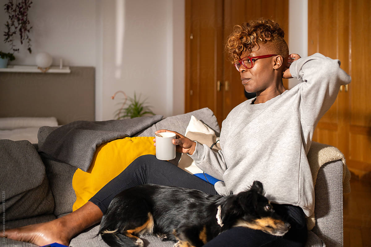 Black Woman Drinking A Cup Of Coffee At Home Next To Her Dog.