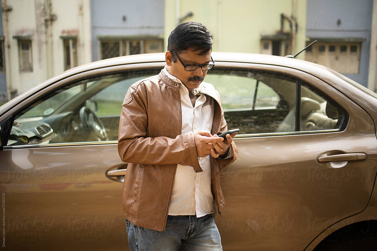 Middle aged Indian man browsing smartphone in front of a luxury car
