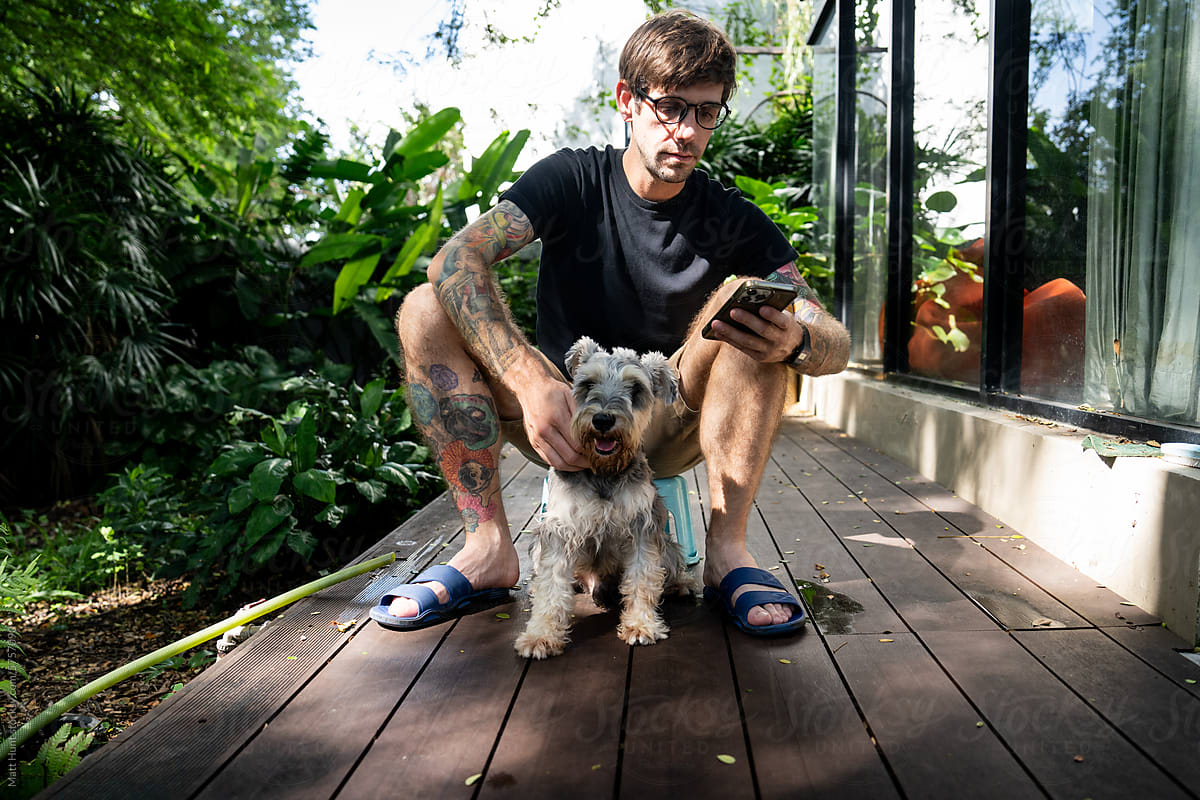 A millennial guy with tattoos checks a phone while holding a schnauzer