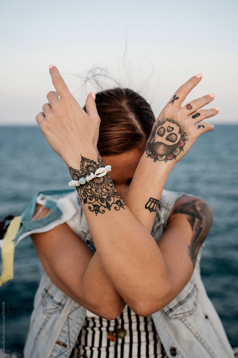 Young stylish woman covering face with tattoed hands in front of sea on pier