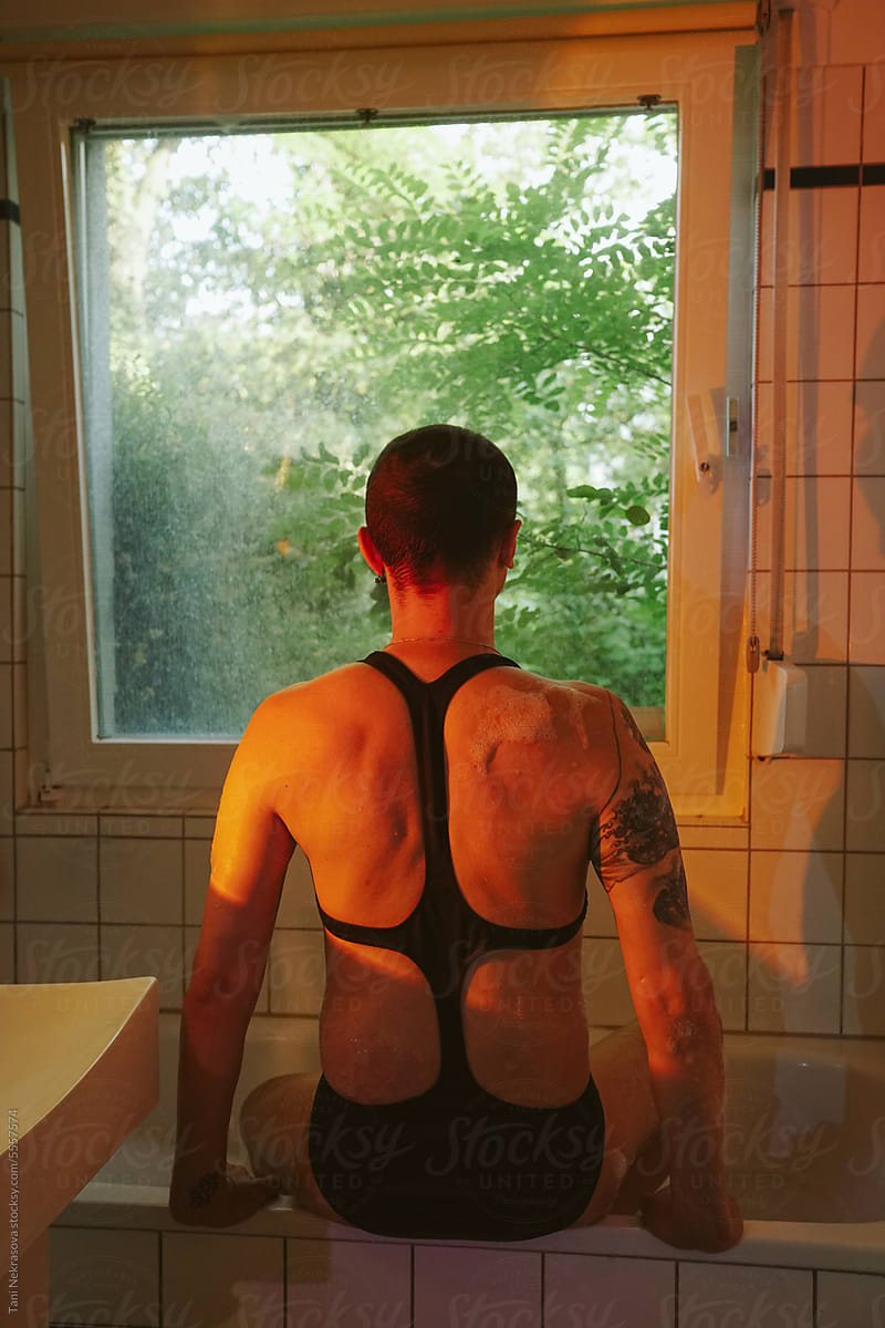 An anonymous man in a swimsuit sits with his back in the bathroom