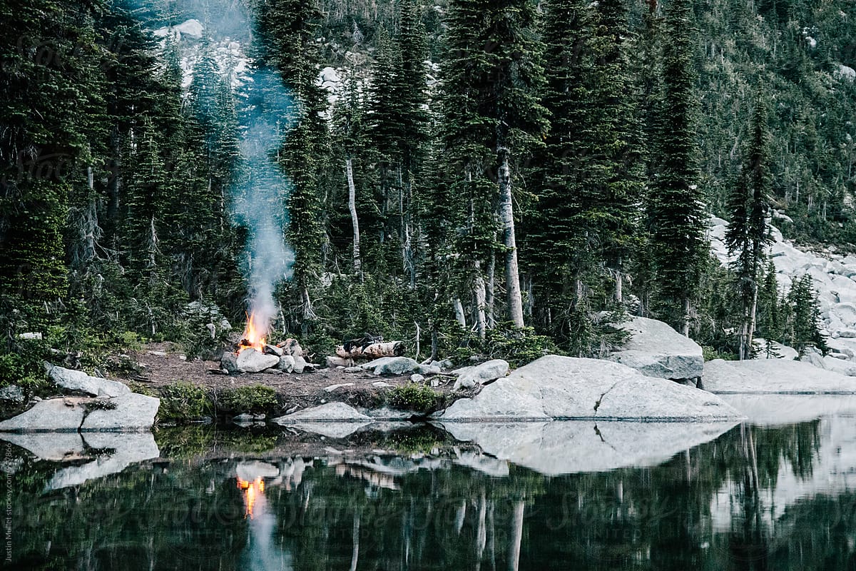 Campfire with plume of smoke beside calm mountain lake.