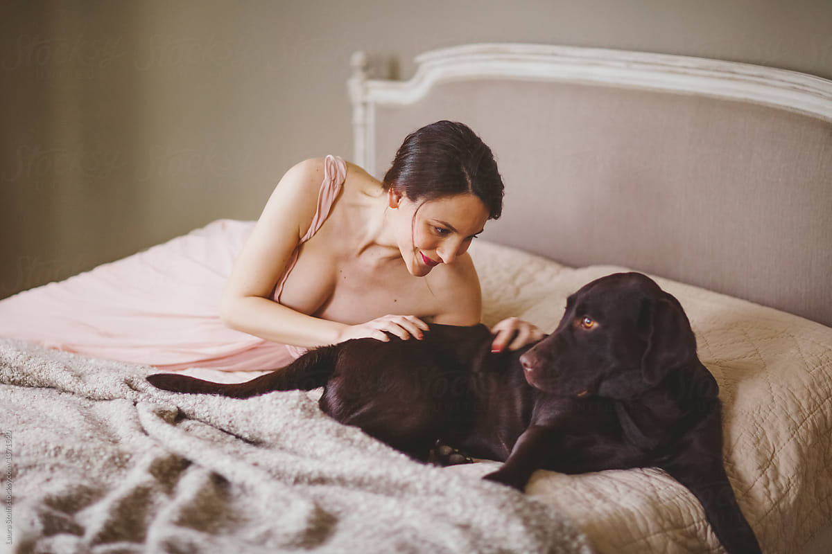 Young woman cuddles her dog on bed in elegant pink dress