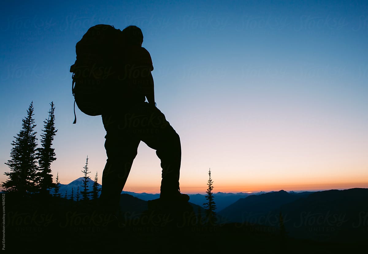 Silhouette Of Male Backpacker Standing On Mountain Summit Taking