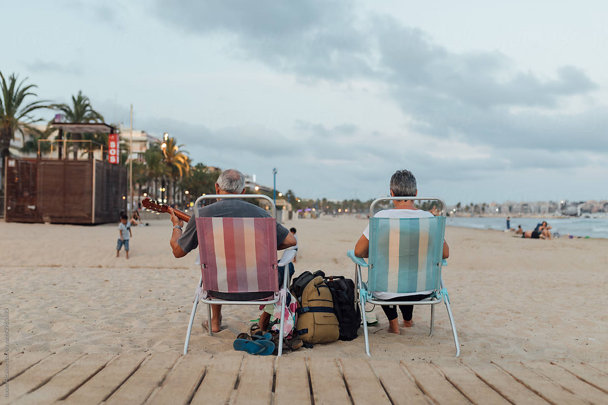 Retired couple playing guitar on a summer afternoon at the beach sitting in striped chairs.