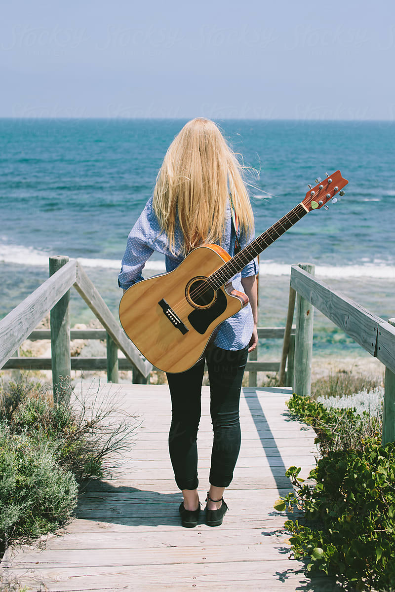 Girl, with a guitar on her back, stands and looks at the ocean.