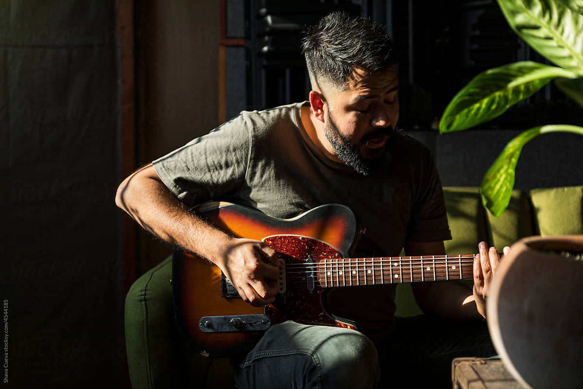 Man with beard singing while playing an electric guitar on a couch
