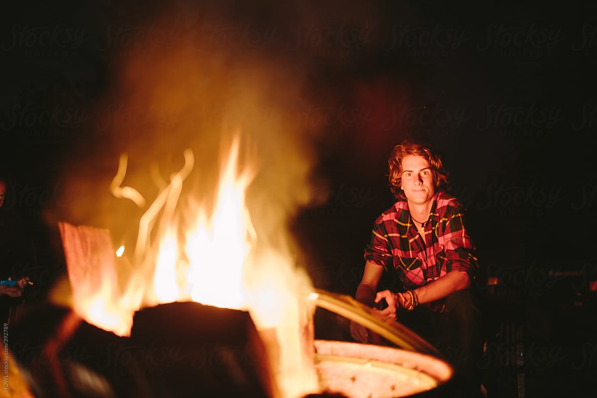 Young boy in flannel sits along side a roaring campfire.
