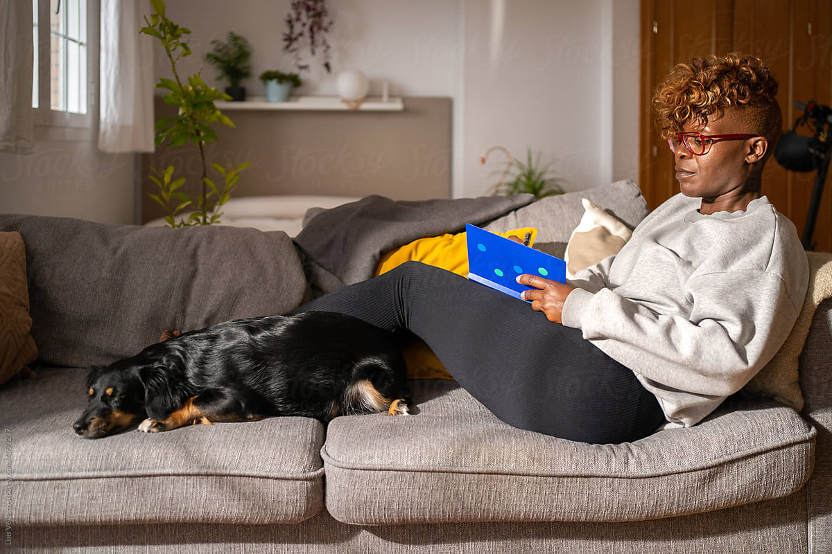 Black Woman With A Book On The Couch Next To Her Dog.