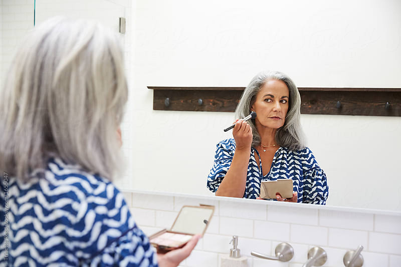 Senior woman with grey hair putting on makeup in bathroom mirror