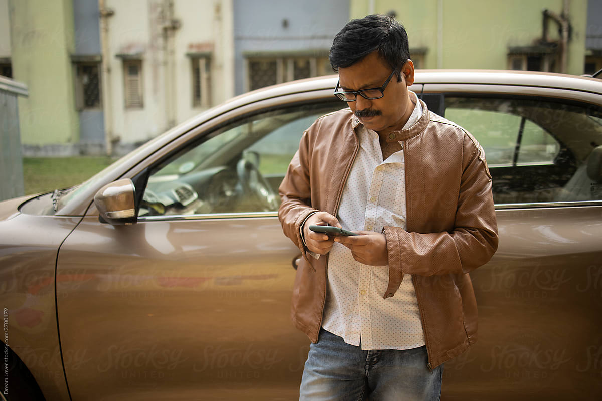 Middle aged Indian man browsing smartphone in front of a luxury car