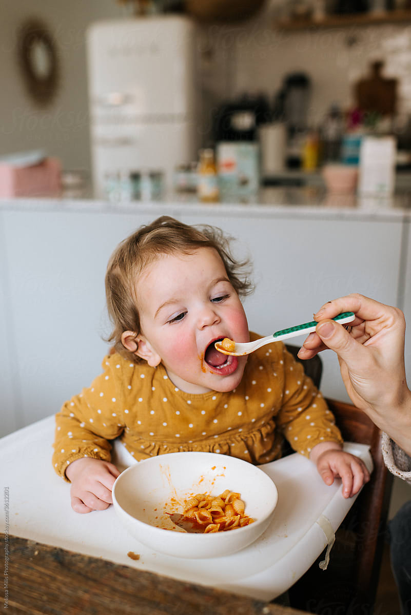 Baby girl with polkadot dress eating pasta in a high chair