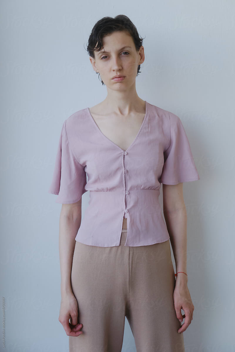 A woman in a pink blouse stands against a white wall.
