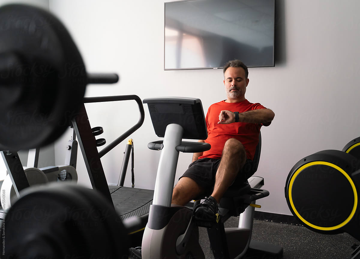 Man At Upright Exercise Bike Looking At Watch
