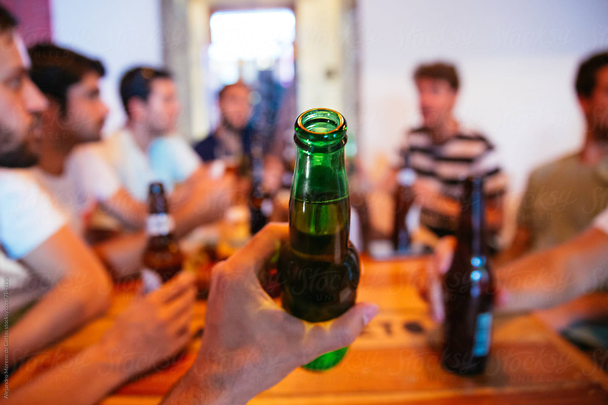 Man hand holding up a full beer bottle in front of camera surrounded by a group of people sitting at a table drinking