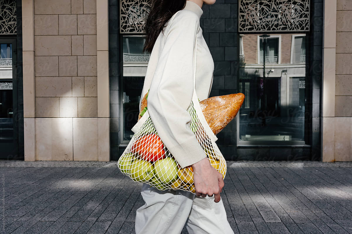 Crop woman walking along street with mesh bag of groceries